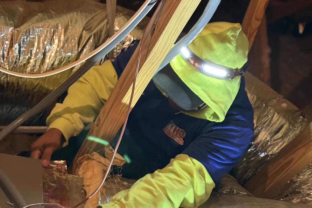 HVAC technician with headlamp inspecting and sealing ductwork in a residential attic with insulation