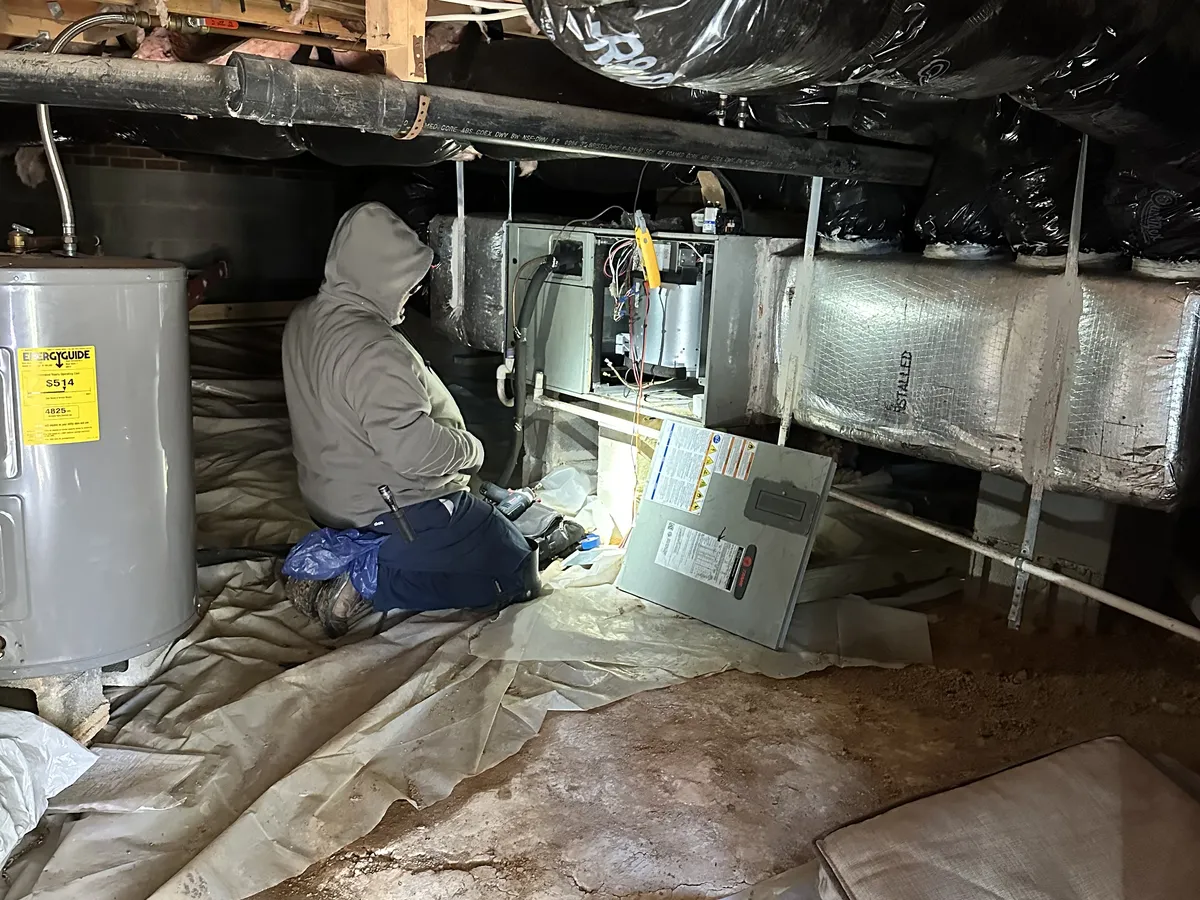HVAC technician performing furnace maintenance in a crawlspace with ductwork and insulation
