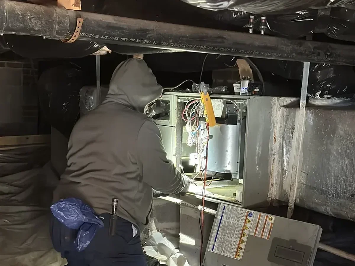 HVAC technician repairing furnace wiring and electrical connections in a crawlspace with exposed ductwork