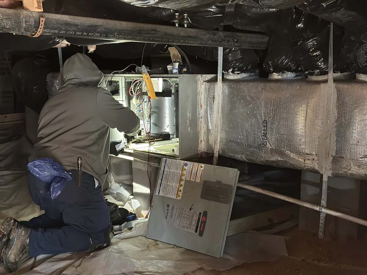 Icy Hot HVAC technician diagnosing and repairing a heating system in a crawlspace with multimeter