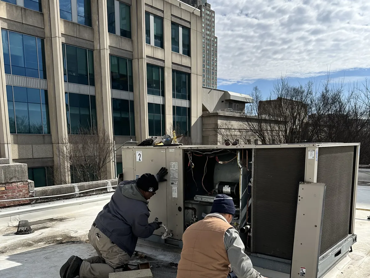 Two HVAC technicians repairing a commercial rooftop package unit with cityscape background