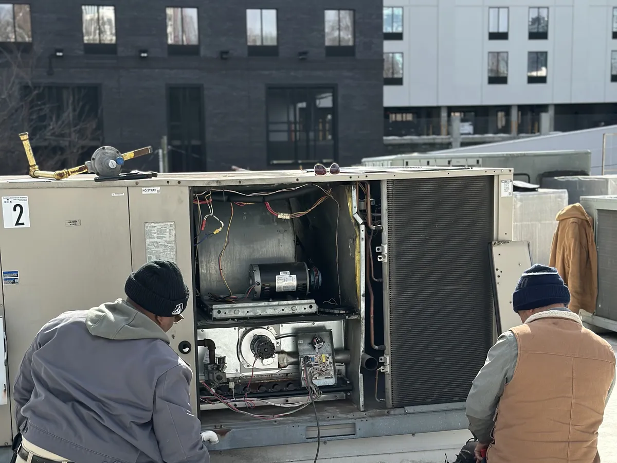 Two HVAC technicians diagnosing and repairing a commercial rooftop package unit with exposed blower motor and wiring